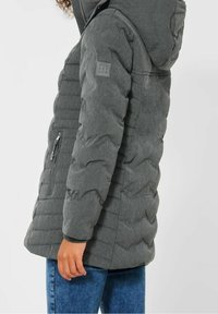 Grå puffjacka med huva, med kviltad struktur och sidofickor med dragkedja. Logotypapplikation på vänster axel. Bärs med blå jeans.