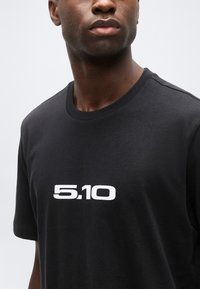 Άνδρας με σκουρόχρωμο δέρμα, φοράει μαύρο T-shirt με λευκό κείμενο "5.10" στο κέντρο του στήθους, μπροστά σε απλό ανοιχτόχρωμο φόντο.