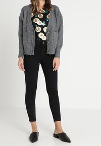 Szary sweter z dzianiny o luźnym kroju, z przednimi kieszeniami naszytymi i zapięciem na guziki, noszony na kwiatowej bluzce i zestawiony z czarnymi skinny jeansami.