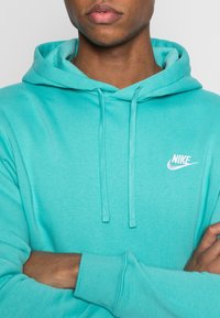 Tyrkysová mikina Nike s kapucí na šňůrku, měkkou texturou a malým bílým logem na hrudi. Dlouhé rukávy a volný střih.