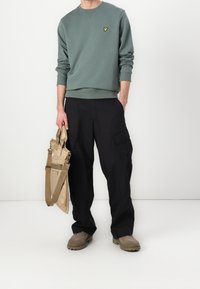 Felpa verde a girocollo con un piccolo logo giallo, pantaloni cargo neri e scarpe beige senza laccio, che tiene una borsa a tracolla beige.