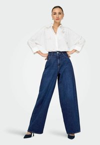 Camicia bianca con ricami floreali, abbinata a jeans a vita alta blu scuro a gamba larga e tacchi a punta blu navy.