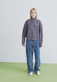 Persoană cu păr blond, purtând un hanorac gri cu fermoar mediu, blugi largi albaștri și adidași albi, stând pe o podea verde, în fața unui perete alb simplu.