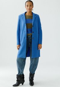 Blauw wollen jasje met revers, zeven knopen en zijzakken, gedragen over een groen-blauw gestreept sweater, gecombineerd met spijkerbroek en zwarte laarzen.