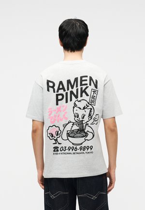 Personne portant un t-shirt gris clair avec des graphismes et des textes sur le thème du ramen en noir et rose, debout de dos à la caméra.