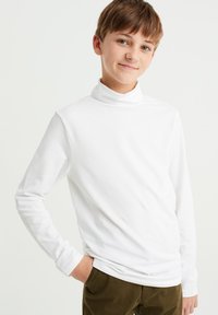 Balta ilgomis rankovėmis turtleneck maikė, pagaminta iš medvilnės, su lygiu paviršiumi ir prigludusia forma, derinama su tamsiai žaliomis kelnėmis.