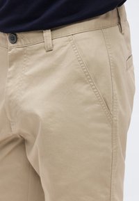 Μπεζ casual παντελόνι με θηλιές ζώνης, κλείσιμο με κουμπί, εμπρόσθιες τσέπες σε γωνία και μαύρο πουκάμισο μαζεμένο στη μέση.