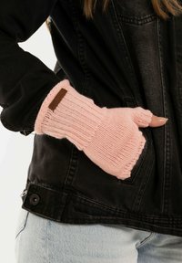 Moufles en tricot rose clair avec un pouce ouvert et une texture côtelée, ornées d'un accent en cuir marron sur le poignet, portées avec une veste noire.