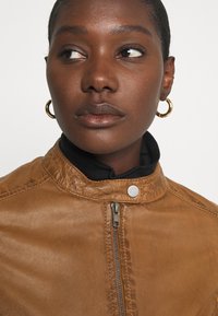 Brown leather jacket with a textured finish, featuring a silver zipper and snap button closure, worn over a black turtleneck. Gold hoop earrings.