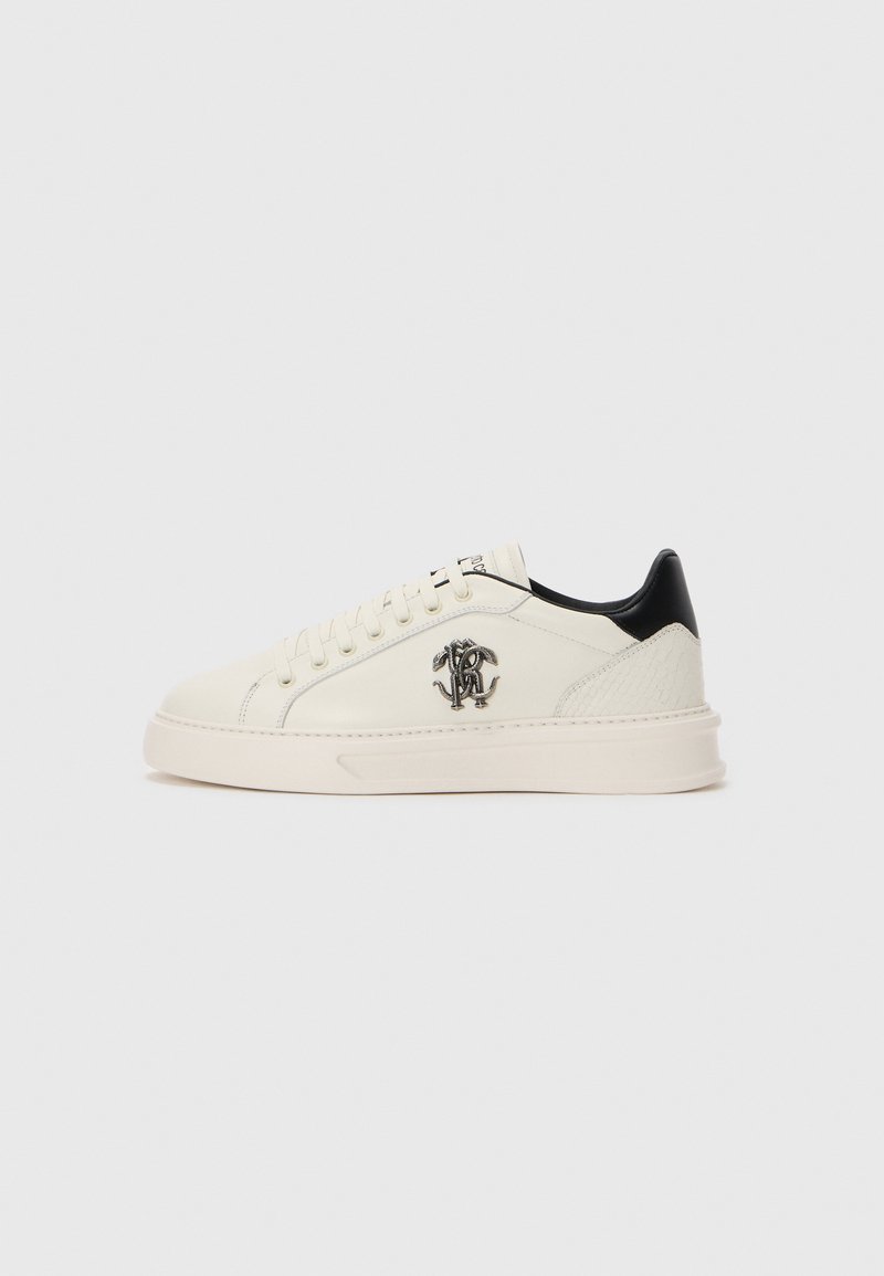 Krémfehér, alacsony szárú sneaker, fűzős elejével, fekete sarok akcentussal, texturált sarokpanellel és fémes monogramos logóval az oldalon.