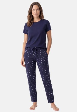 Kvinde står barfodet iført en navyblå kortærmet top og navyblå pyjamasbukser med hvide hjerte mønstre.