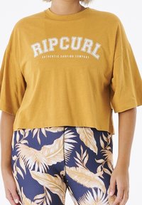 Musztardowa krótka koszulka z krótkim rękawem z logo "RIPCURL" w białym kolorze, o luźnym kroju, połączona z niebieskimi legginsami w kwiatowy wzór.