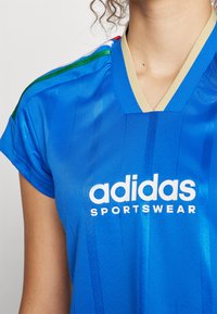 Zils sporta krekls, kas izgatavots no gludas auduma, ar horizontālām svītrām un bēša krāsas apkakli. Uz tā ir baltais "adidas SPORTSWEAR" logo.