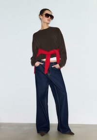 Rjava ohlapna jopa z rdečo vezalko v pasu, kombinirana s širokimi temno modrimi kavbojkami in rjavimi čevlji z ostrim vrhom. Oblečena velika sončna očala.