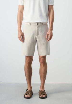 Hombre con pantalones cortos beige hasta la rodilla, camisa blanca de manga corta y sandalias marrones, de pie frente a un fondo claro y liso.