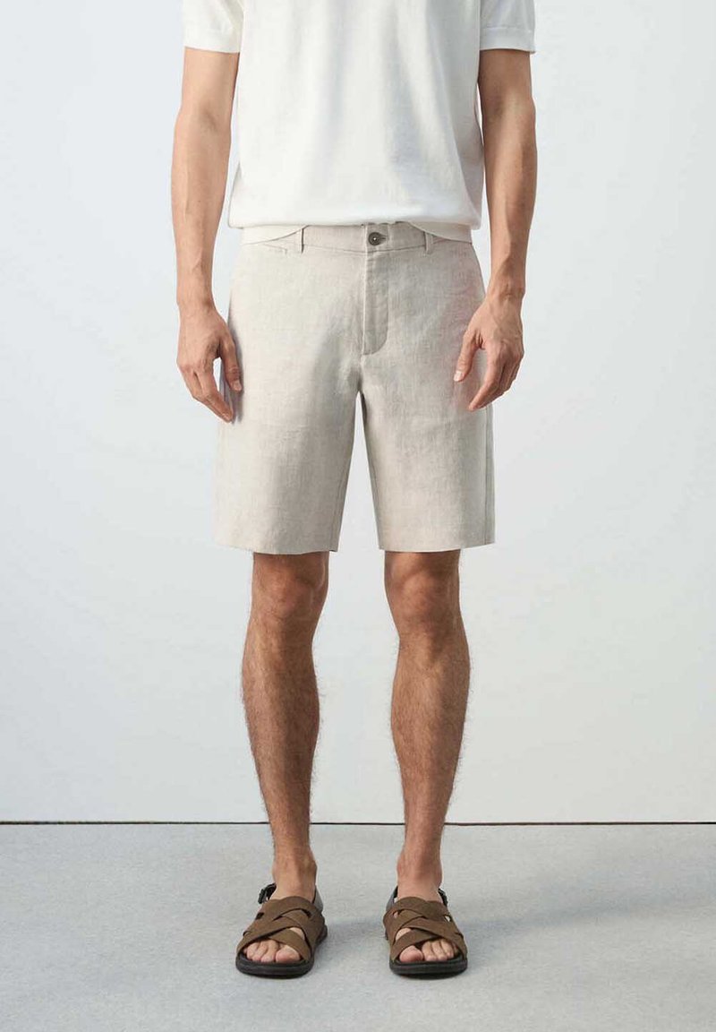 Homme portant un short beige au niveau des genoux, une chemise blanche à manches courtes et des sandales marron, debout devant un fond clair uni.