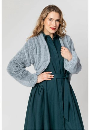 Kobieta z długimi blond włosami nosi turkusową sukienkę z wiązaniem w talii i jasnoszary krótki sweter z dzianiny, pozując z jedną ręką na biodrze.