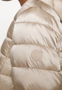 Beige puffer-takki, jossa on tikkaukset ja sileä, kiiltävä pinta. Sisältää pyöreän "Save the Duck" -logon olkapäällä.