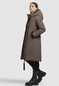 Langer brauner Puffer-Mantel mit Chevron-Muster, durchgehendem Reißverschluss und Kapuze. Schwarze Akzente an den Taschen und einem Lanyard-Tag. Getragen mit schwarzen Kampfstiefeln.