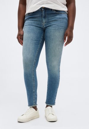 Persona con pantalones vaqueros ajustados azul claro con dobladillos deshilachados y zapatillas blancas, de pie frente a un fondo liso.