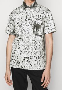 Camicia a maniche corte bianca con pattern a macchie nere, dotata di tasca con stampa di serpente e etichetta del logo. Collo button-down, vestibilità comoda.