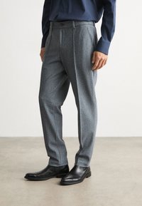 Muž v šedých kostkovaných kalhotách, černých kožených botách a tmavě modré košili s dlouhým rukávem stojí na jednobarevném podlaze.