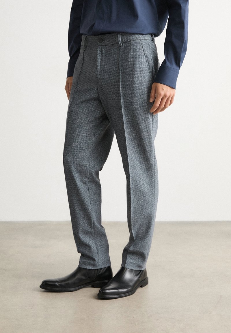 Muž v šedých kostkovaných kalhotách, černých kožených botách a tmavě modré košili s dlouhým rukávem stojí na jednobarevném podlaze.