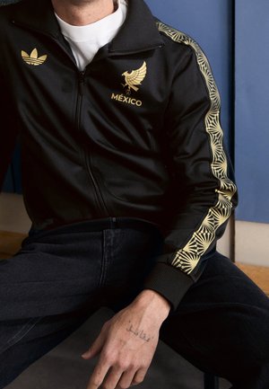 Persoon draagt een zwart trainingsjack met gouden tekst "México", adelaarsembleem, Adidas-logo en gouden patroonstrepen langs de mouwen.