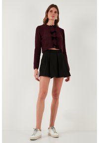 Žena v krátkém burgundském cardiganovém svetru s černými mašlemi, černé plisované sukni a bílých teniskách stojící na čistém bílém pozadí.