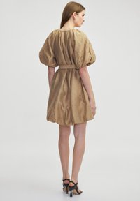 Korte, beige jurk met pofmouwen en een getailleerde taille. Heeft een textuurstof en een vloeiende rok. Zwarte hoge hakken maken de look compleet.