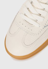 Close-up of beige textured leather sneaker toe with white laces and gum rubber sole on white background.