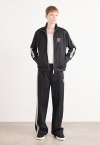 Zwarte trainingsjack en broek met witte gestreepte accenten. Bevat een rits en logo op de borst. Materiaal lijkt glad en sportief.