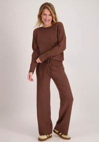 Bruine gebreide trui met een ronde hals en lange mouwen, gecombineerd met bijpassende wijde broekspijpen. De outfit heeft een zachte textuur en een trekkoorddetail.