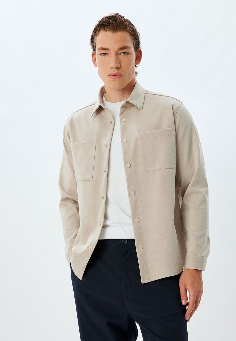 Camisa beige de botones con dos bolsillos en el pecho, tejido texturizado y un cuello. Llevada sobre una camiseta blanca, combinada con pantalones oscuros.