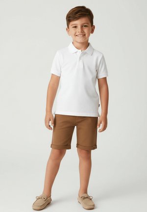 Młody chłopiec stoi i się uśmiecha, ubrany w białą koszulkę polo, brązowe podwinięte szorty oraz beżowe mokasyny, na jednolitym tle.