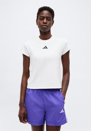 Mladá osoba nosiaca biele tielko bez rukávov a fialové športové šortky s logami Adidas, stojaca na jednoduchom pozadí.