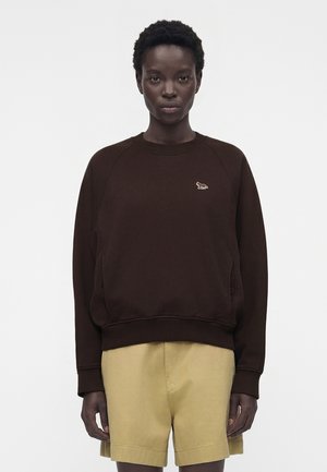 Person wearing a dark brown sweatshirt with a small animal logo on the chest and beige knee-length shorts, standing against a plain background.