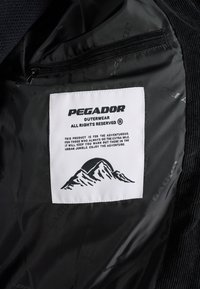 Giacca nera con interno caratterizzato da un'etichetta bianca con la scritta "PEGADOR" e un grafico di montagna. Include una tasca con cerniera e una texture del tessuto liscia.