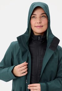 Giacca esterna impermeabile di colore teal con cappuccio, dotata di isolamento nero sotto, tasche con zip e dettagli in Velcro per chiusure sicure.