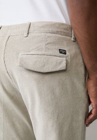Pantalon en velours côtelé beige avec une texture à côtes verticales, doté d'une poche arrière à rabat et d'une petite étiquette de marque noire sur la poche.