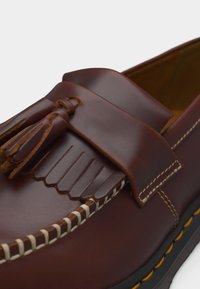 Καφέ δερμάτινο μοκασίνι με φουντίτσες, λευκή αντίθεση ραφής κατά μήκος της άκρης και μαύρη σόλα με κίτρινη λεπτομέρεια ραφής.