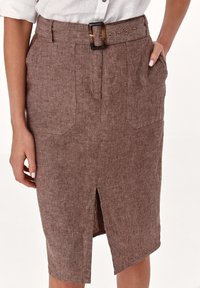 Nainen, jolla on polveen saakka ulottuva ruskea teksturoitu hame, jossa on edessä halkio, tilkkupocketit ja kilpikonnakuosiset vyö, yhdistetty valkoiseen käänteellä varustettuun paitaan.