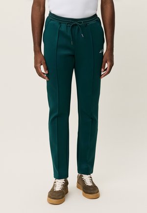 Grønne polyesterbukser med snøre i taljen, med sidelommer og en tætsiddende pasform, kombineret med brune sneakers med hvide snørebånd.