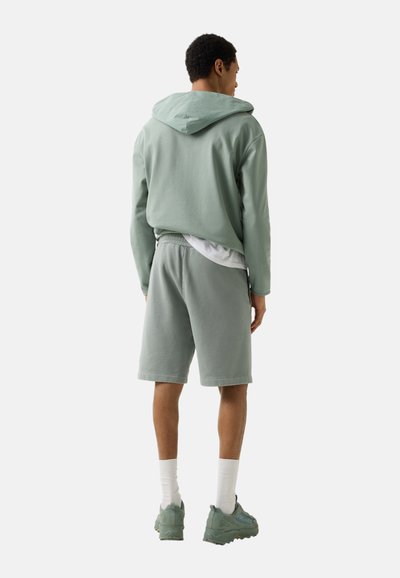 Homme portant un sweat à capuche vert clair, un short assorti, des chaussettes blanches et des baskets vertes, debout de dos sur un fond uni.