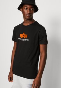 Juodas medvilninis marškinėlis, turintis oranžinę ir baltą grafiką su užrašu "Alpha Industries" ir žvaigždės dizainu. Apvalus kaklas ir trumpi rankovės.