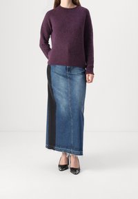 Femme portant un pull violet, une jupe longue en denim bicolore avec un ourlet effiloché, et des escarpins noirs à bout pointu, debout devant un fond blanc.