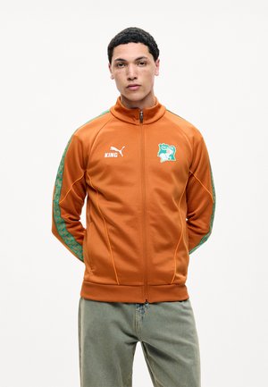 Ung mand iført en orange Puma lynlåstrøje med grønne mønstrede striber og Elfenbenskystens fodboldlogo, stående med hænderne bag ryggen.