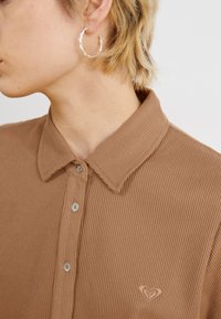 Polo shirt marron à côtes avec une finition texturée, col et trois boutons. Présente un petit logo sur le côté gauche de la poitrine.