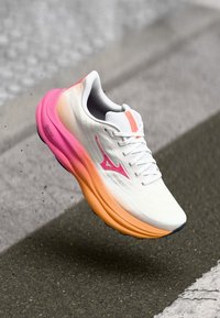 White athletic shoe with a textured upper, pink and orange gradient sole, and contrasting pink logo on the side. Lightweight and breathable design.