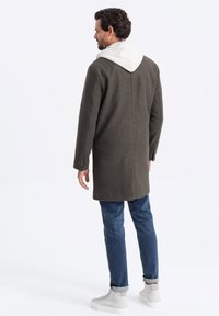 Manteau long, vert foncé avec une capuche gris clair, éventuel simple et deux boutons sur les manches, associé à un jean bleu et des baskets gris clair.